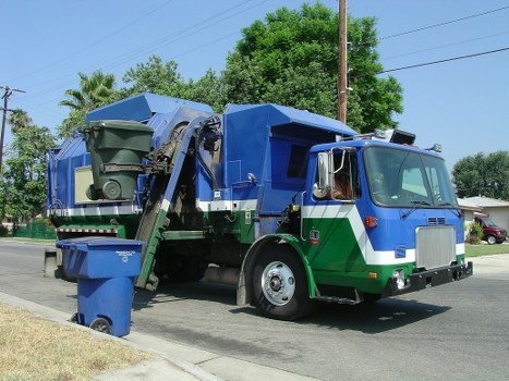 A large, blue and green rubbish removal truck parked on a suburban street with trees and utility poles in the background. The truck features a side-loading mechanism with a refuse container attached at the rear, partially lifted for waste collection. The vehicle has a prominent cab with a curved windshield and side mirrors, and the body is painted in shades of blue with green accents on the lower section. The street has a paved road and a grassy verge, typical of residential areas in Chertsey, nearby the postcode area. The scene is captured in daylight, with clear skies and calm surroundings, indicating an active waste collection operation by House Clearance Chertsey, specializing in domestic rubbish disposal and clearance services.