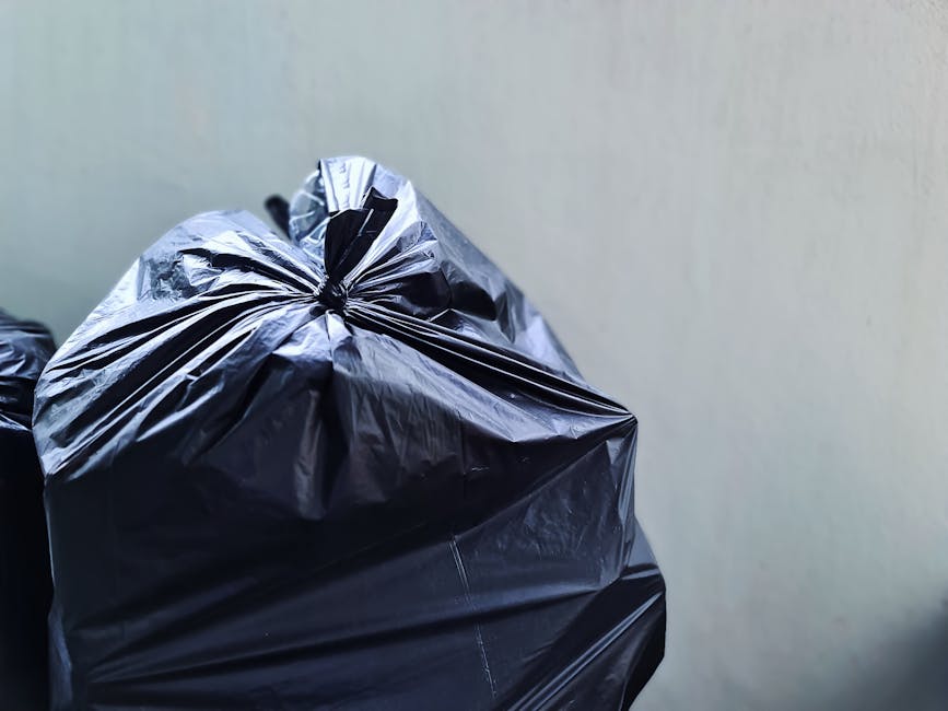 Close-up of a large black plastic rubbish bag filled with waste, tightly knotted at the top, resting against a plain light-colored wall. The bag's glossy surface reflects light, emphasizing its crinkl