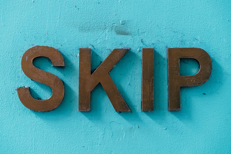 Close-up view of three three-dimensional brown metal letters spelling 'SKIP' mounted on a weathered, light blue painted wall. The letters have a matte finish, with some slight rusting and surface irre