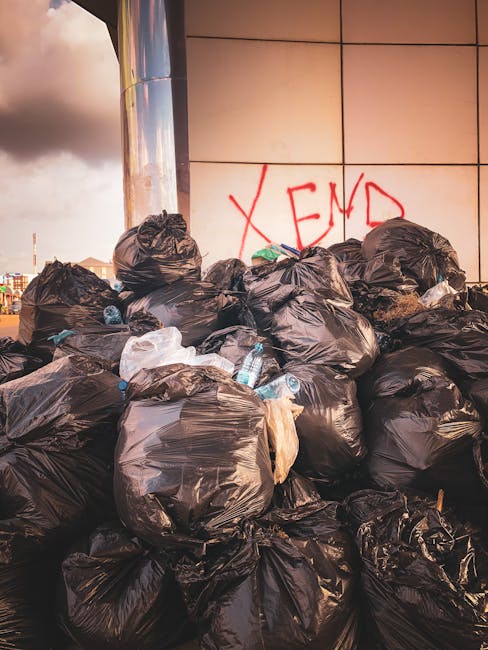 A row of large, wheeled wheelie bins lined up along a pavement section adjacent to a brick wall. The bins are made of durable plastic, with textured, ribbed lids and smooth sides, and each is painted 